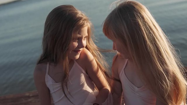 Two girls are sitting on the beach and smiling. Wooden pier Slow motion. In the background sea sunset.