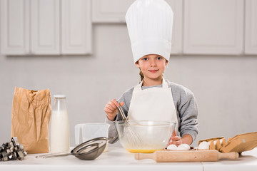 smiling child in chef hat whisking eggs in bowl at table in kitchen