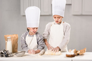 children in aprons and chef hats making dough with rolling pin at table in kitchen