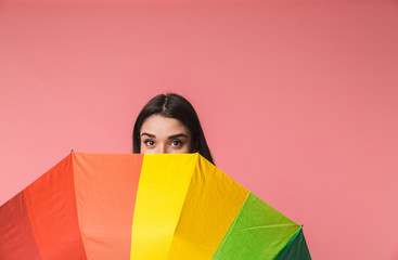 Emotional young woman posing isolated over pink background holding rainbow umbrella.