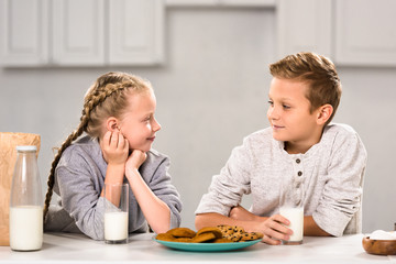 smiling kids looking at each other and sitting at table with cookies and milk in kitchen