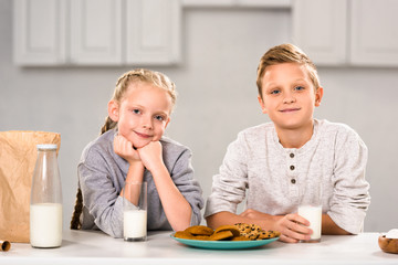 Obraz na płótnie Canvas happy brother and sister looking at camera near table with milk and cookies