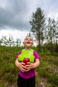 Outdoor portrait of a happy smiling little boy wearing a bib and holding a red soda can. Forest nature scene.