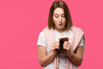 shocked young woman looking into empty wallet isolated on pink