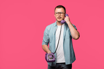 serious young man in eyeglasses talking by vintage telephone isolated on pink
