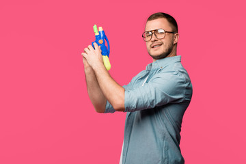 young man in eyeglasses holding toy gun and smiling at camera isolated on pink