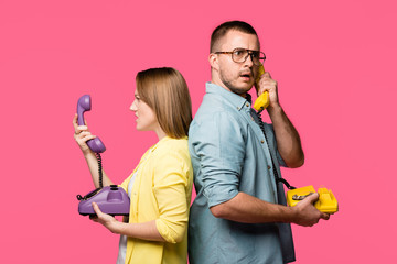 side view of angry young couple standing back to back and talking by rotary phones isolated on pink
