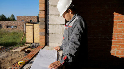 Side view of man in hardhat standing at window in house under construction looking at paper draft