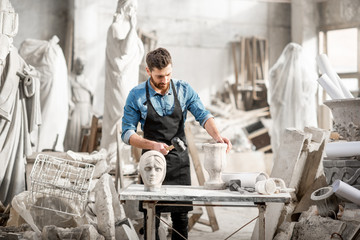 Portrait of a handsome sculptor in blue t-shirt and apron working with stone sculptures on the table at the old atmospheric studio