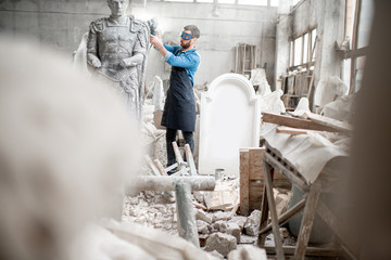 Sculptor working with big stone figure grinding with electirc grinder in the old atmospheric studio with different sculptures on the background