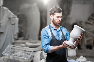 Portrait of a handsome sculptor in blue t-shirt and apron holding old vase in the studio with...