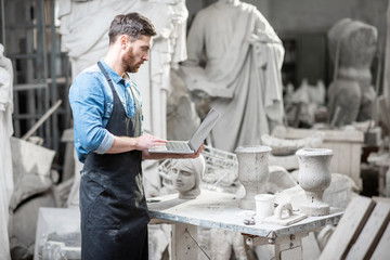 Handsome sculptor working with laptop in the atmospheric studio with old sculptures on the...