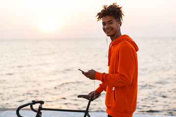 Young african guy outdoors on the beach using mobile phone listening music walking with bicycle.