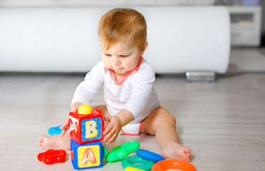 Adorable baby girl playing with educational toys in nursery. Happy healthy child having fun with colorful different toys at home. Kid trying to build plastic pyramid and using blocks with letters