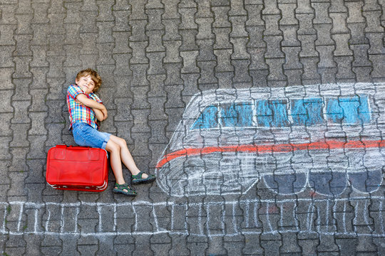 Little kid boy having fun with fast train picture drawing with colorful chalks on asphalt. Child painting with chalk and crayon and going on vacations. Schoolkid sitting on suitcase. Travel concept.