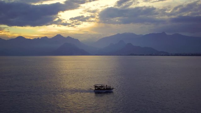 Tourists travelling by small ship in scenic marine background. Beautiful sunset sunny sea water, mountains, cloudy fluffy sky, sailing yacht and  Antalya city landscape. Real time 4k video footage.