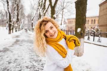 Blond curly playful hipster woman on street, ljumping, enjoying winter smiling at snowy park. Model wearing white sweater, yelow knitted hat, sarf, gloves. City lifestyle concept.