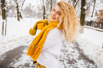Blond curly playful hipster woman on street, ljumping, enjoying winter smiling at snowy park. Model wearing white sweater, yelow knitted hat, sarf, gloves. City lifestyle concept.