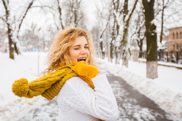 Blond curly playful hipster woman on street, ljumping, enjoying winter smiling at snowy park. Model wearing white sweater, yelow knitted hat, sarf, gloves. City lifestyle concept.