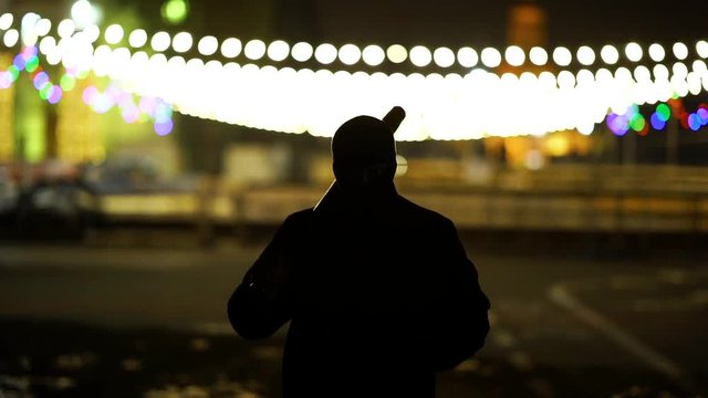 A man dressed in dark clothes and a balaclava goes with a bat in an abandoned park. There is an early winter outside and garlands hang around. His face is not visible in backlight.