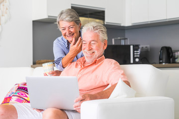 aged senior caucasiann couple sit down on couch using an internet device connected laptop together having fun and smiling - retired life for man and woman old people at home enjoying lifestyle 