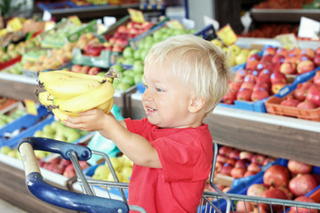 Cute toddler boy holds bananas in his hands while sitting in shopping cart inside supermarket