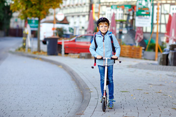 Active school kid boy in safety helmet riding with his scooter in the city with backpack on sunny day. Happy child in colorful clothes biking on way to school.