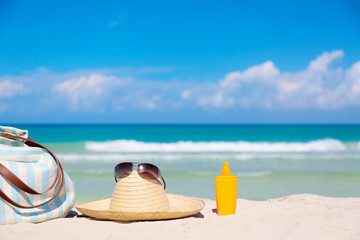 Beach accessories on sand for summer vacation concept. Bag, straw hat with sunglasses and sunscreen lotion bottle. White sand with amazing ocean and blue sky in the background.