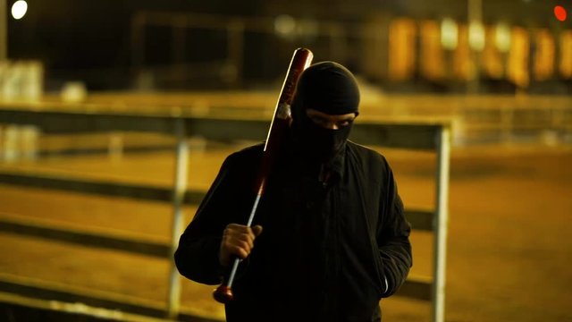 A man dressed in dark clothes and a balaclava goes with a bat in an abandoned park. There is an early winter outside and garlands hang around. His face is not visible in backlight. He is set up to fig