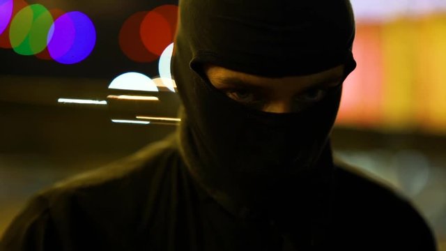 A man dressed in dark clothes and a balaclava goes with a bat in an abandoned park. There is an early winter outside and garlands hang around. His face is not visible in backlight. He is set up to fig