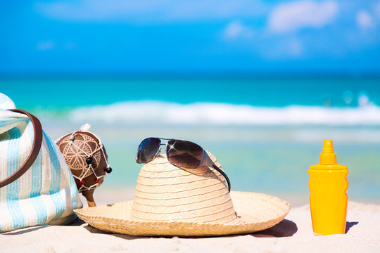Beach accessories on sand for summer vacation concept. Bag, maracas, straw hat with sunglasses and sunscreen lotion bottle. White sand with amazing ocean and blue sky in the background.