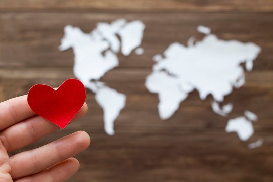 Hand holding red heart and world map cutted from white paper on the wooden background. Love concept. Close-up, soft focus, copy space, mock up