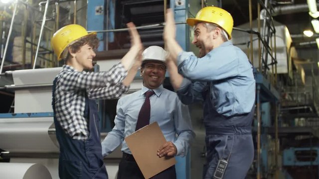 Happy Latin American supervisor and Caucasian male and female workers in overalls and hardhats smiling, showing thumbs up, giving high five and embracing while celebrating success in factory