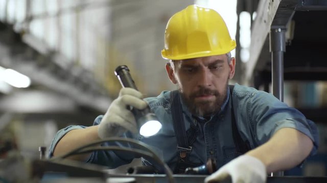 Tilt down of bearded Caucasian male technician in uniform and protective hardhat inspecting industrial equipment with flashlight while working in factory