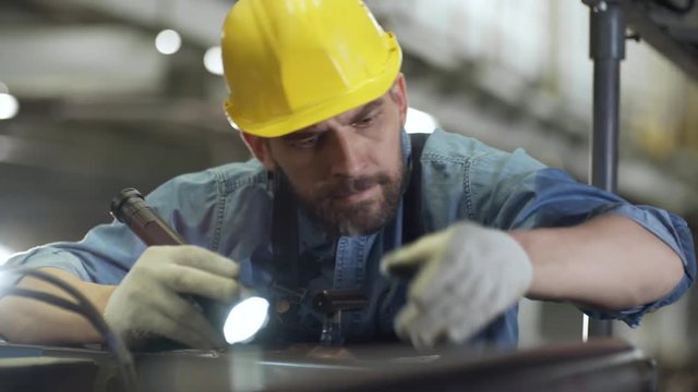Middle aged Caucasian male technician in uniform and protective hardhat using flashlight while repairing industrial equipment in factory
