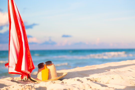 Beach accessories on sand for summer vacation concept. Umbrella, straw hat with sunglasses and sunscreen lotion bottle. White sand with amazing ocean and blue sky in the background.