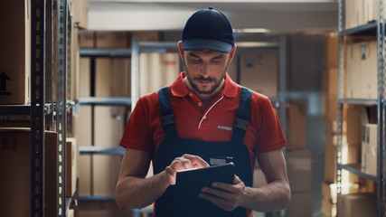 Warehouse Worker Uses Digital Tablet For Checking Stock, On the Shelves Standing Cardboard Boxes.