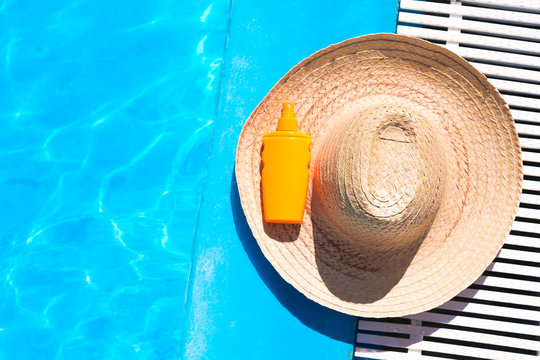Straw hat with sunscreen lotion bottle near the surface of blue clear swimming pool with free space. Summer vacation concept.