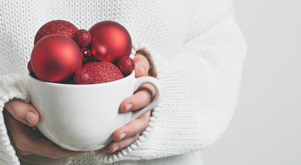 Woman in warm white sweater holding white mug in hands with christmas balls with effect snowfall. ...