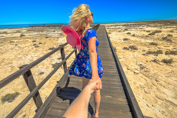 Follow me. Woman in hat holding hand of her friend and walking on wooden path at Stromatolites Hamelin Pool. Marine Reserve, Shark Bay, Western Australia. Girl enjoys of Australian travel destination.
