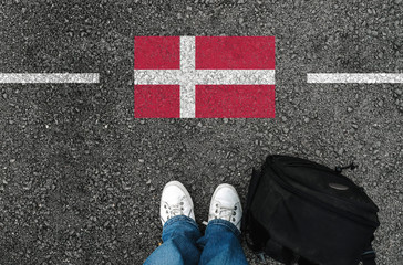 a man with a shoes and backpack is standing on asphalt next to flag of Denmark and border.