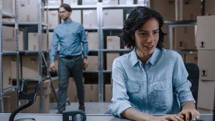 Female Inventory Manager Sitting at Her Desk and Using Personal Computer, Worker Puts Packages on the Designated Shelf. In the Background Rows of Cardboard Boxes with Products Ready For Shipment.