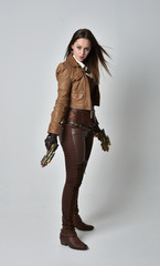 full length portrait of brunette  girl wearing brown leather steampunk outfit. standing pose, holding a gun, on grey studio background. © faestock