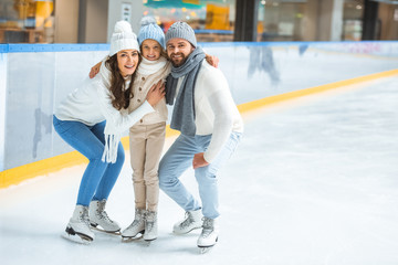 cheerful parents and daughter in sweaters looking at camera on skating rink