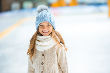 portrait of happy kid in knitted hat looking at camera on skating rink