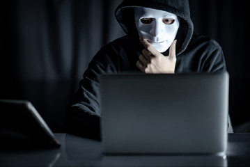 Mystery male hoodie hacker holding white mask looking at laptop computer. Anonymous social masking....