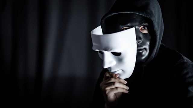 Mystery hoody man wearing black mask holding white mask. Anonymous social masking. Major depressive disorder or bipolar disorder. Halloween concept