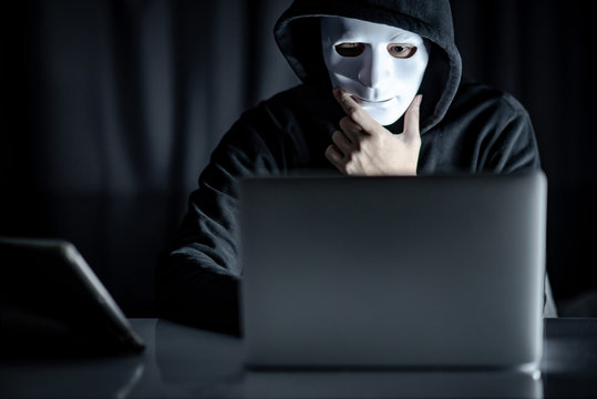 Mystery male hoodie hacker holding white mask looking at laptop computer. Anonymous social masking. Ransomware cyber attack or internet security concepts