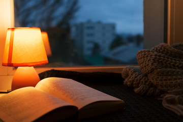 Wool scarf, small table lamp and old vintage book on window sill. Cozy home in the dusk