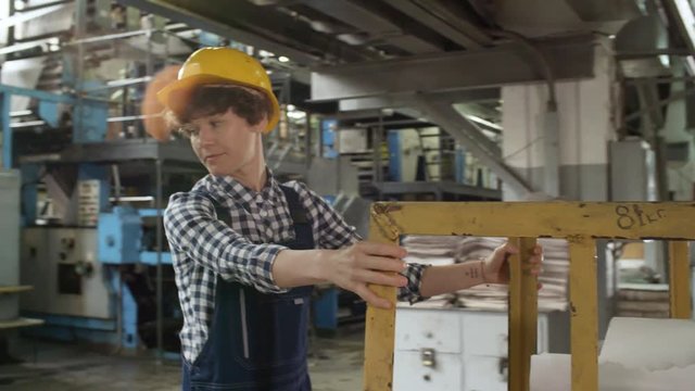 Young female worker in overall and hardhat pulling cart with paper reels while working in printing plant
